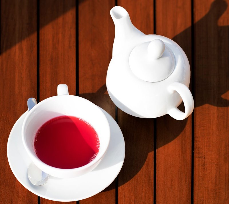Overhead shot of a white teacup and teapot with hibiscus tea on a wooden table.