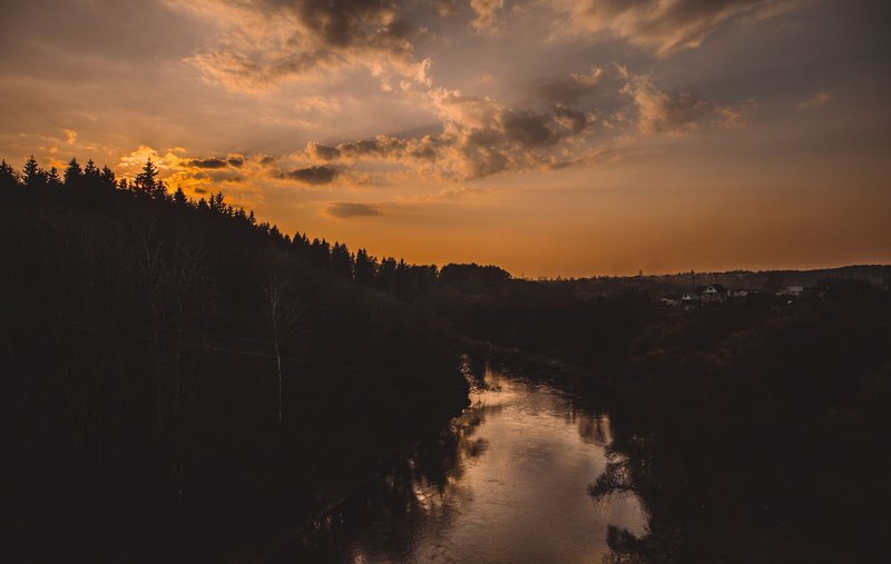 A serene sunset over a forested river with an orange sky reflecting on the water, creating a peaceful atmosphere.