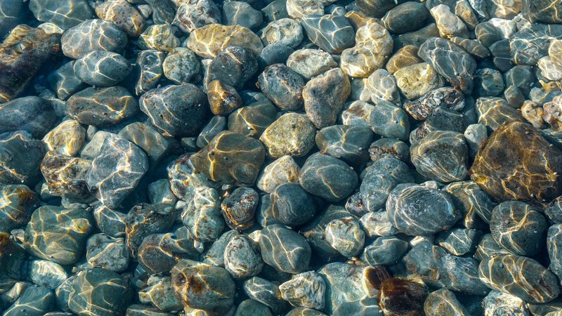 Sunlight refracts through clear water revealing smooth pebbles beneath.