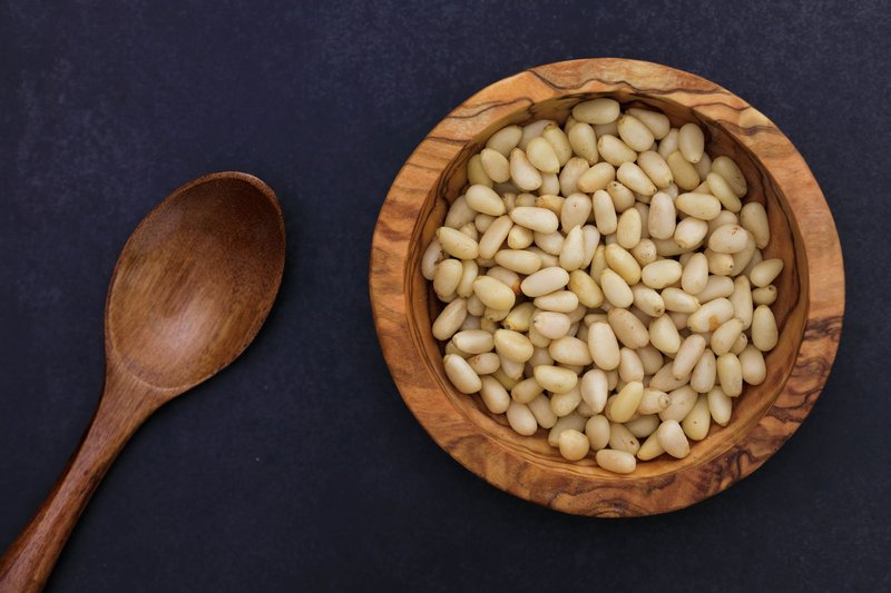 A close-up of pine nuts in a wooden bowl with a rustic spoon on a dark background.