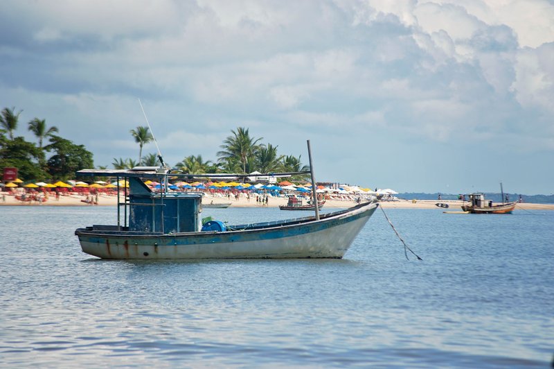 A peaceful scene of a fishing boat floating near a tropical beach with palm trees and sun umbrellas.
