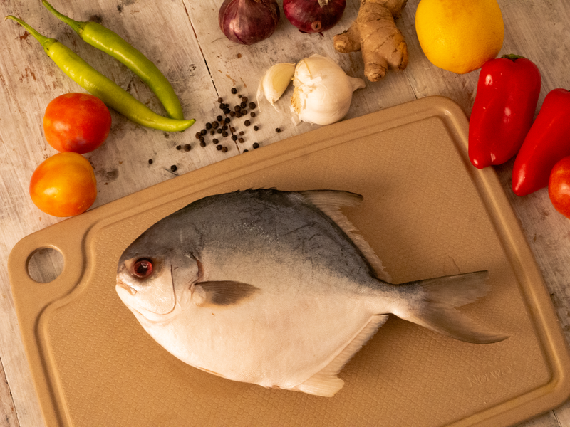 A fresh pomfret fish with vibrant vegetables and spices on a wooden cutting board.